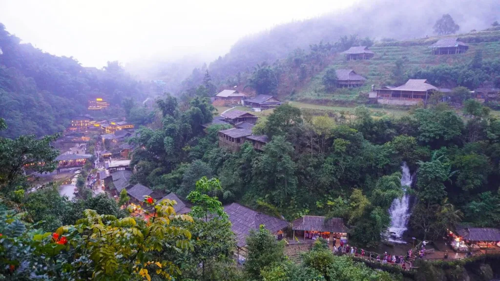 Vista panorámica de un valle verde con casas de madera iluminadas, niebla y una cascada en el norte de Vietnam
