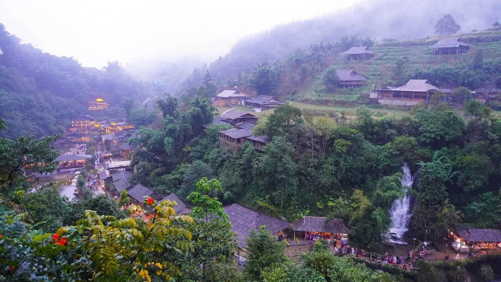 Vista panorámica de un valle verde con casas de madera iluminadas, niebla y una cascada en el norte de Vietnam